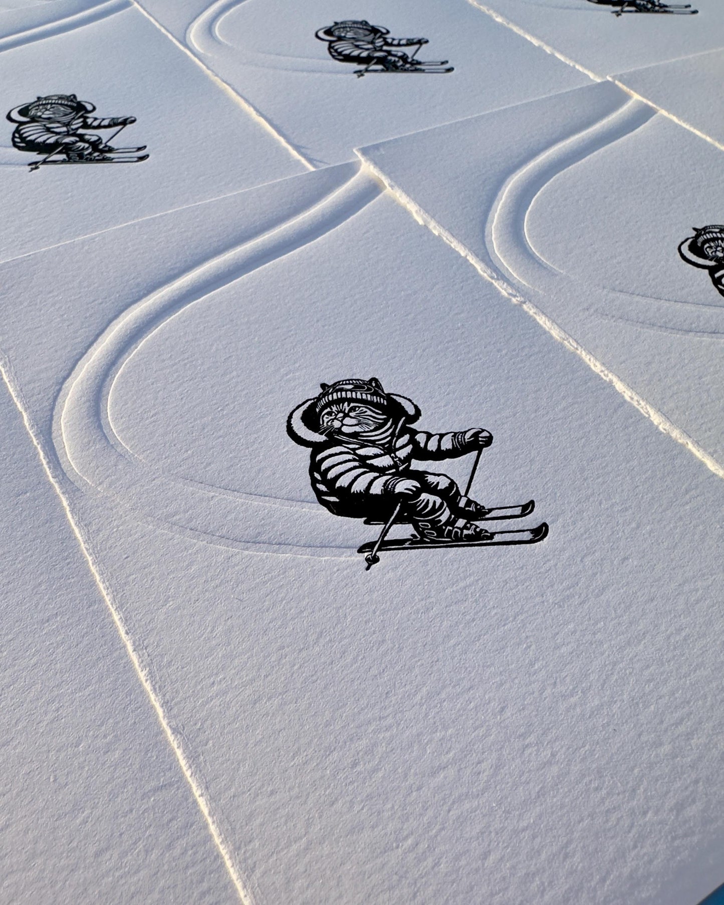 A cat dressed in ski gear descending a snowy mountain, this black and white linocut print perfect for ski lovers and cat lovers alike. The freshly made tracks in the snow are embossed into the paper.